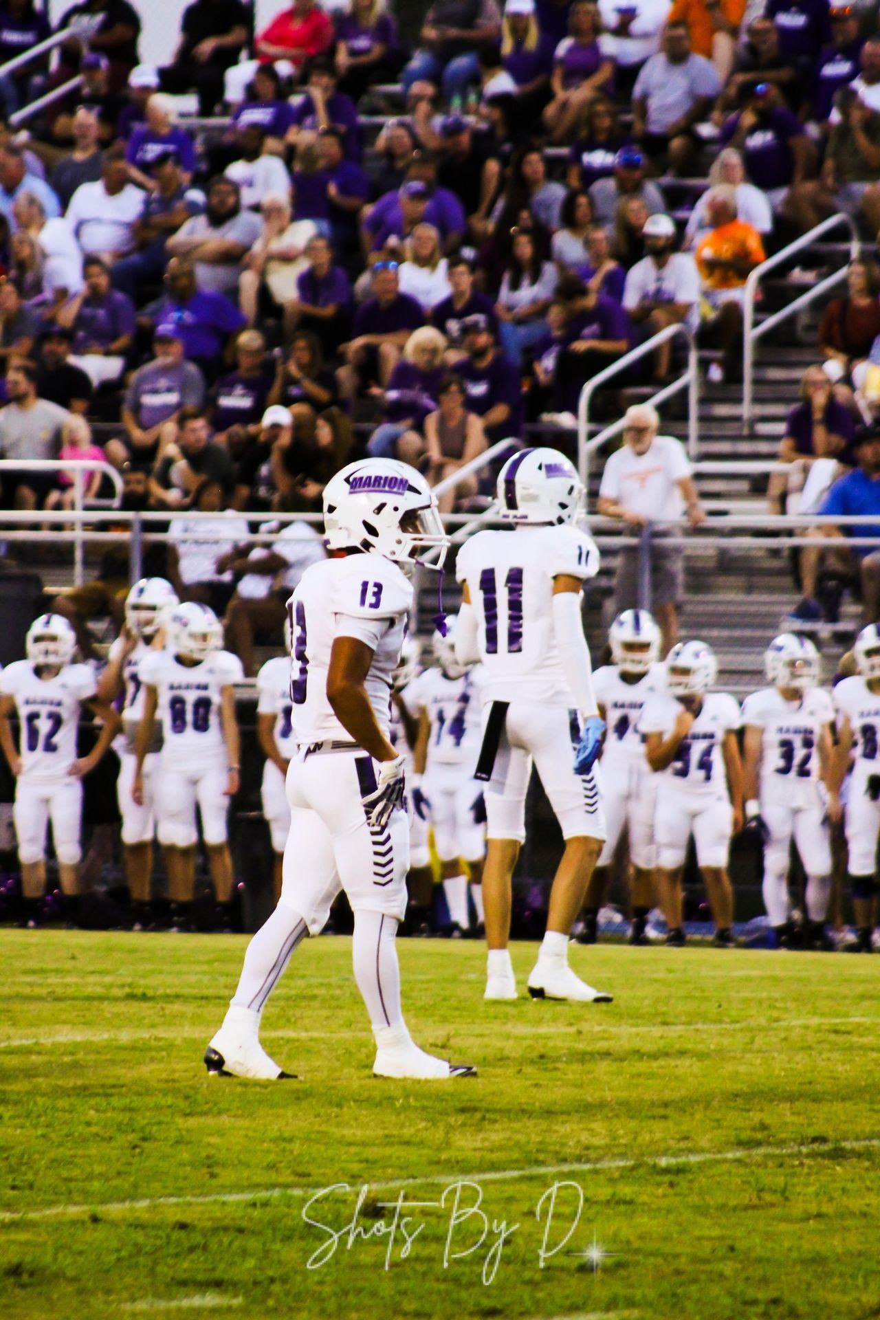 High school football players in white uniforms on the field with a crowd of spectators in the background.