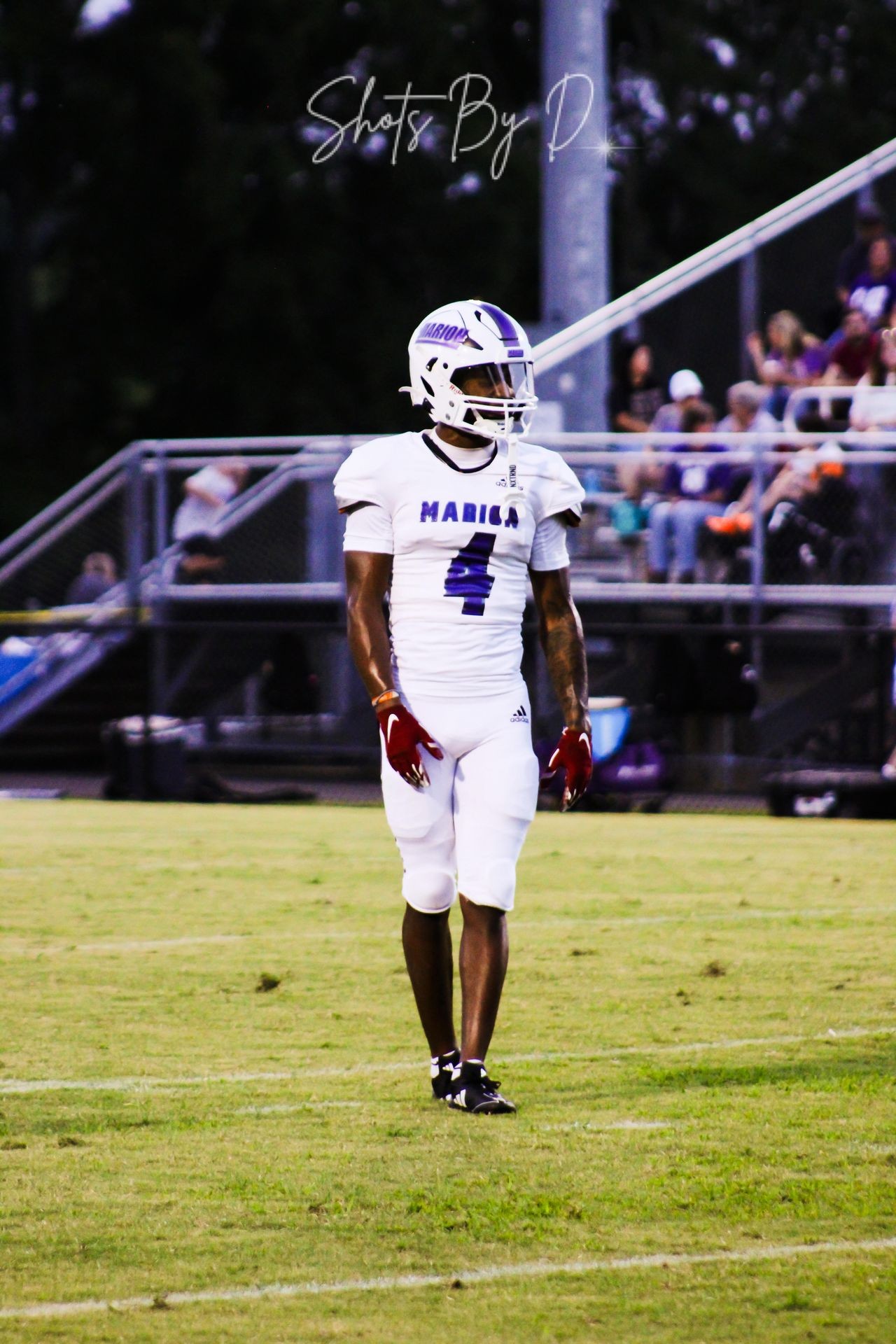 Football player in white uniform and helmet standing on the field with spectators in the background.