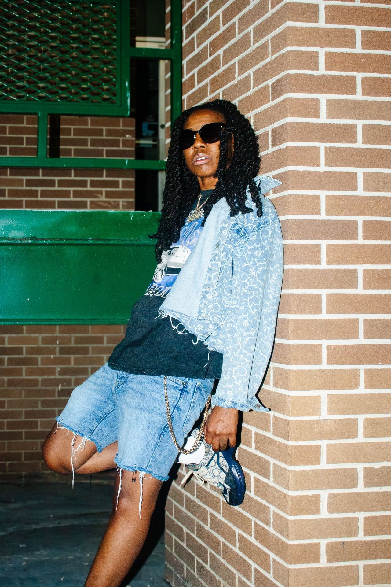 Person with long curly hair wearing sunglasses, denim jacket, and shorts leaning against a brick wall.