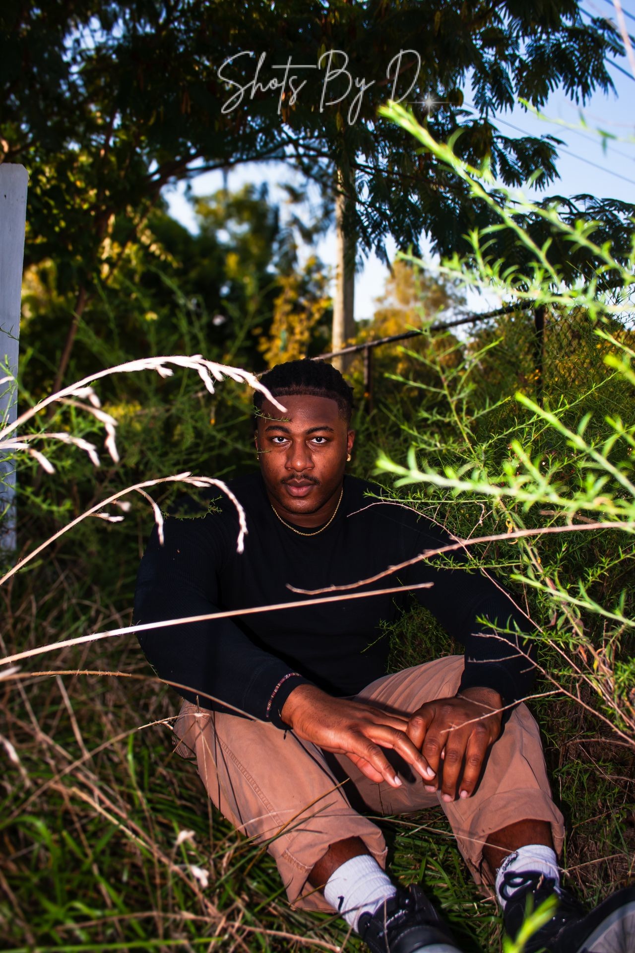 Person sitting on the ground surrounded by green foliage, wearing a black shirt and beige pants.